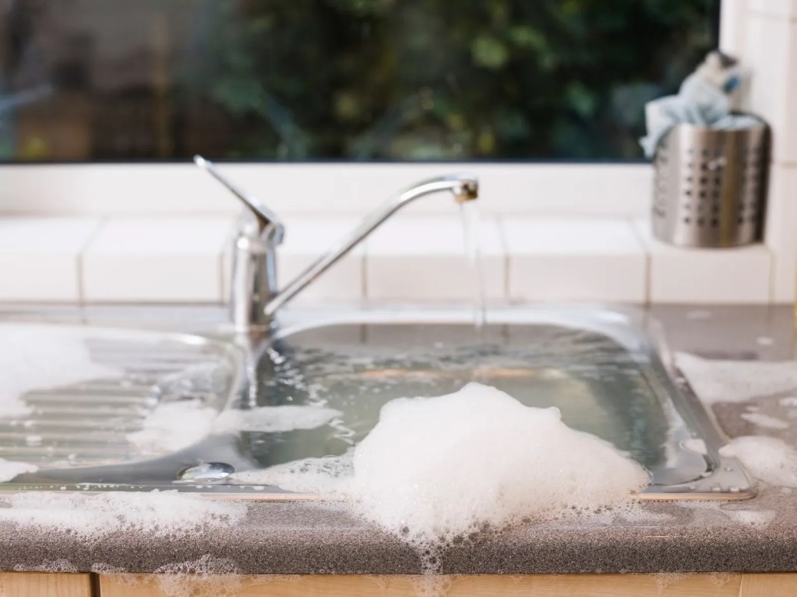 Kitchen sink with running water and soapy foam bubbles on countertop near window during cleaning.