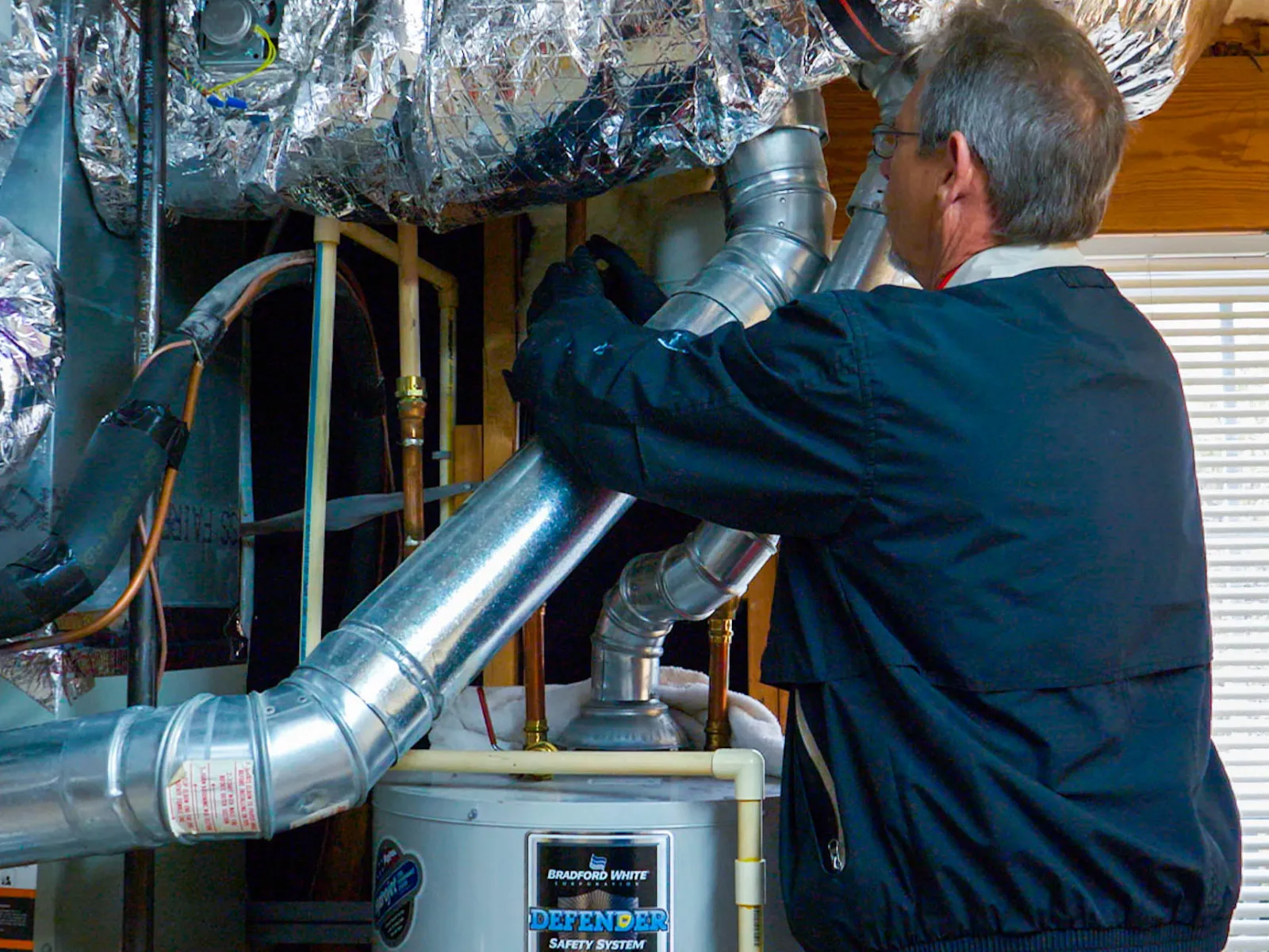 Technician servicing Bradford White Defender water heater with ventilation pipes and insulated ductwork.