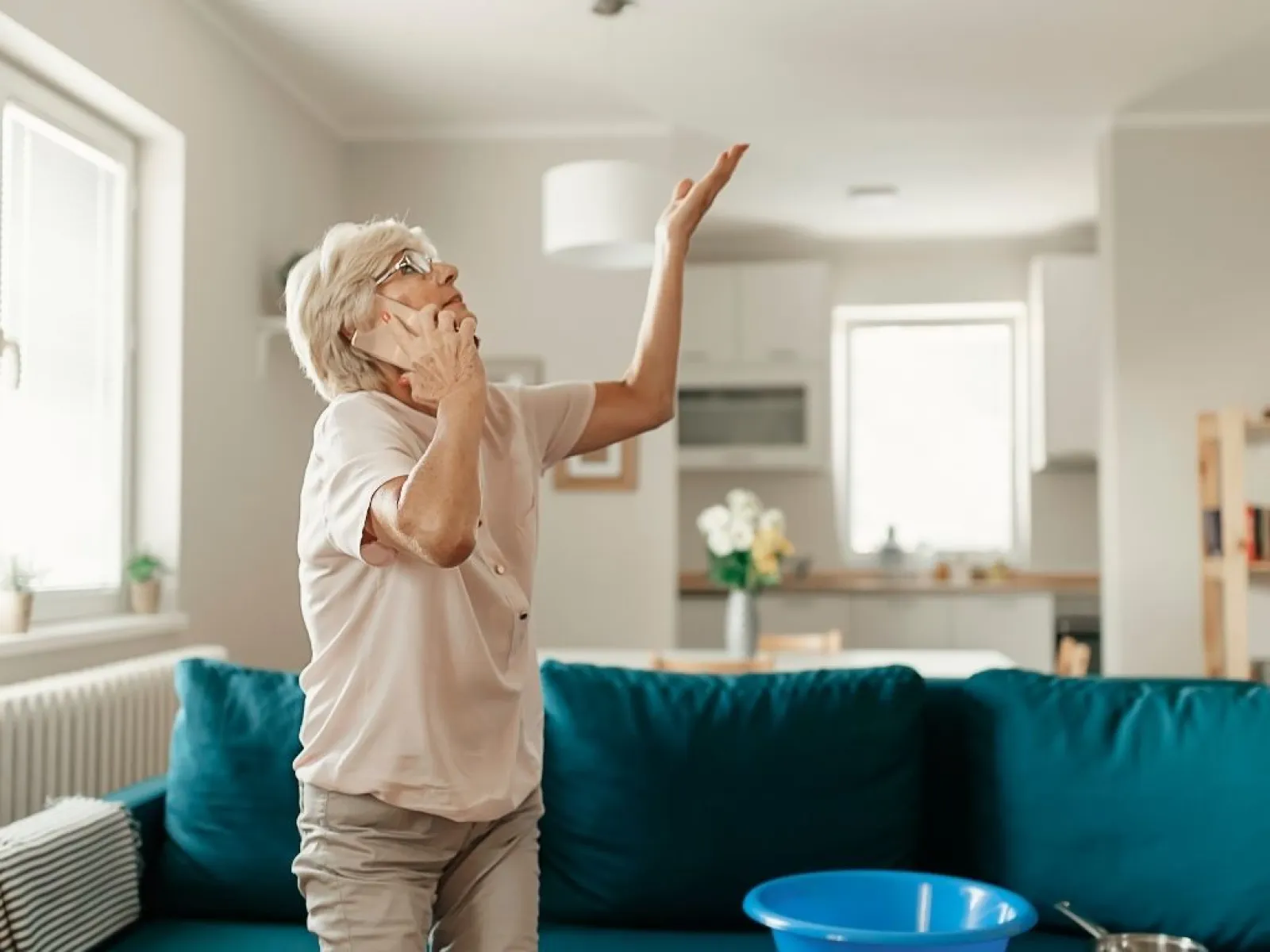 Elderly woman holding phone and raising hand toward ceiling in a living room with blue couch and kitchen background.