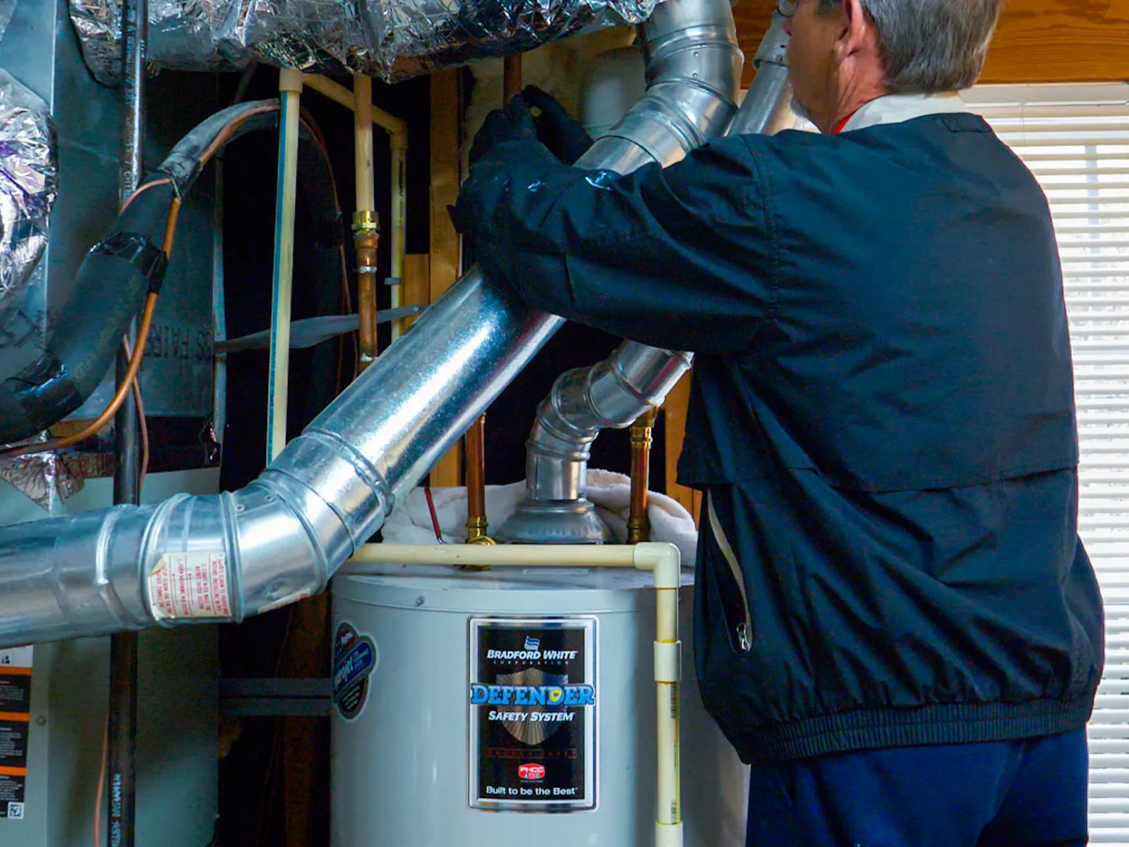 Technician adjusting plumbing expansion tank above a Bradford White water heater in a home utility space.