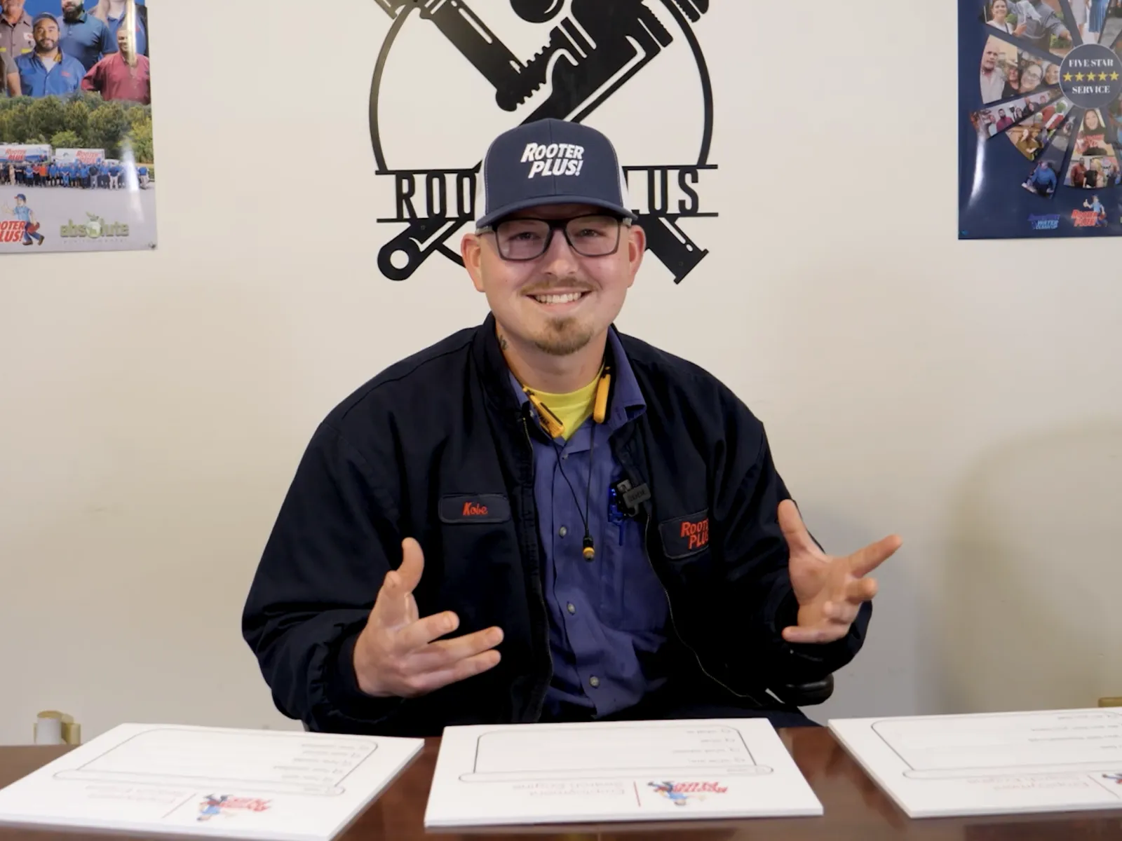 A smiling man in a cap sits at a table with papers, showcasing a friendly demeanor in an office setting.