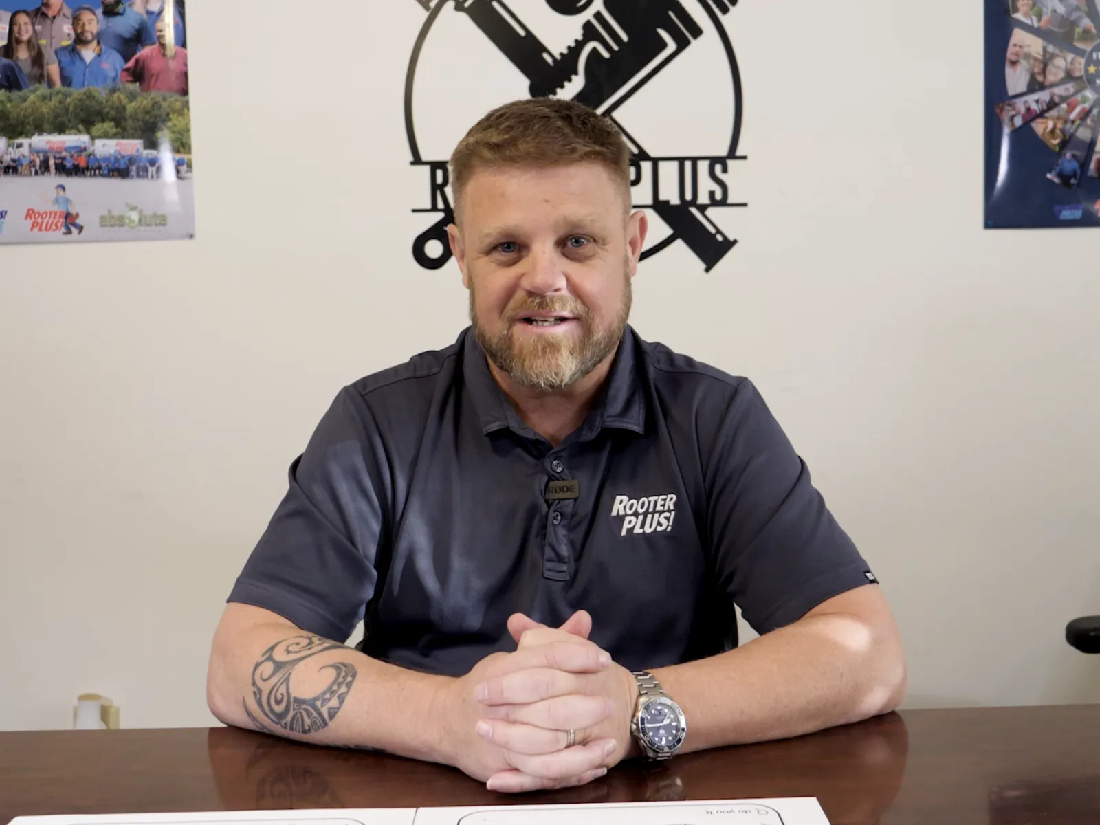 Man in a polo shirt smiling at a desk with a logo in the background, exuding professionalism.
