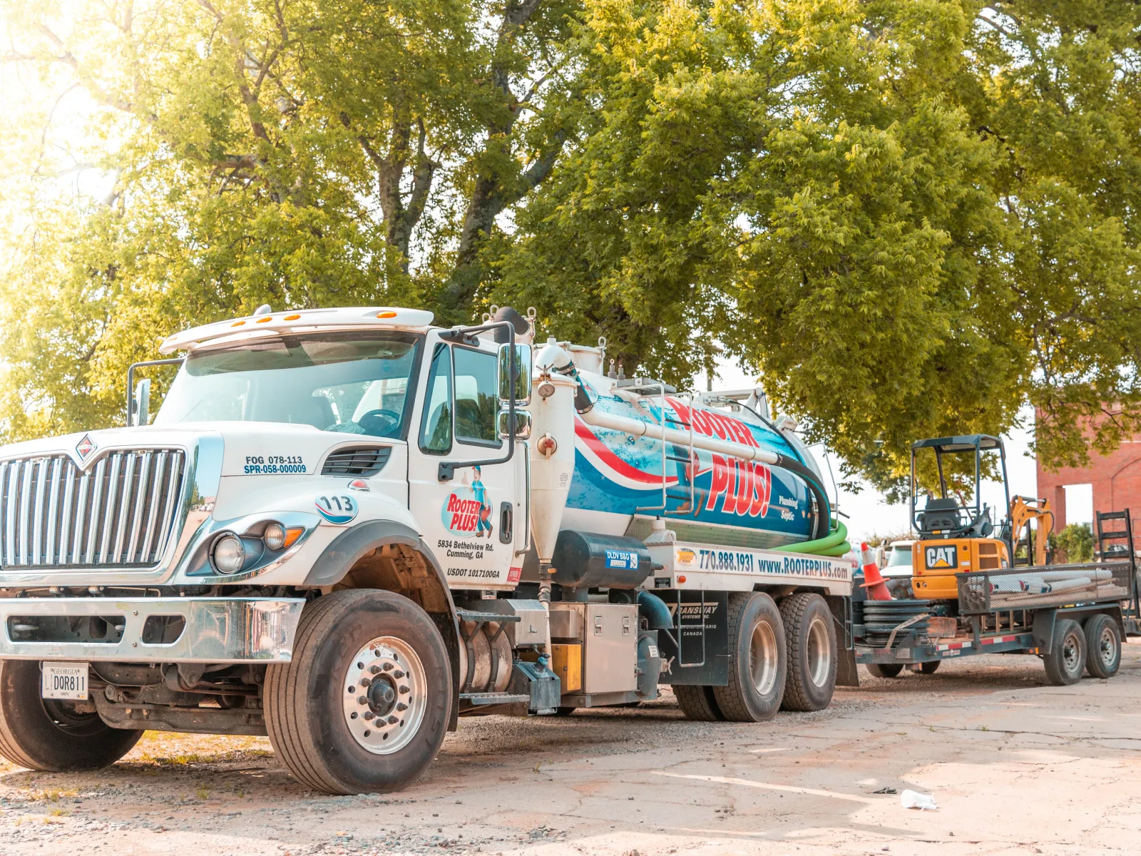 White Rooter Plus septic service truck parked beside large tree with trailer carrying construction equipment.