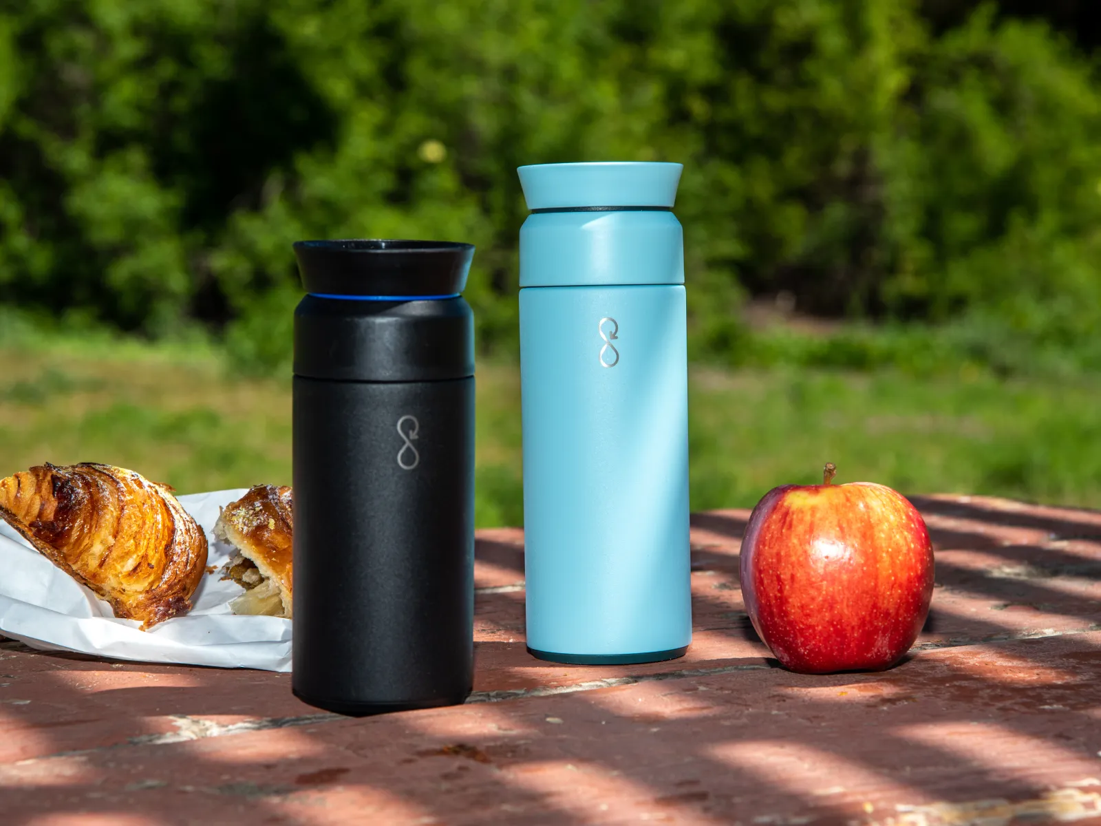 Black and blue travel mugs on a picnic table with croissants and a red apple in natural outdoor setting.