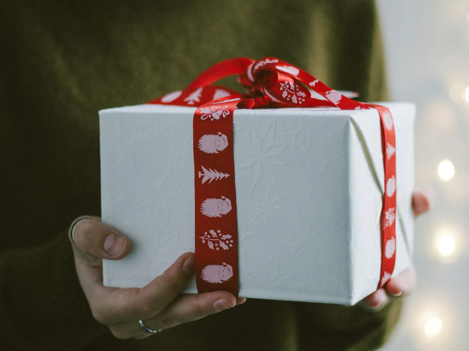 Hands holding a white gift box wrapped with a red ribbon featuring hedgehogs and festive designs.