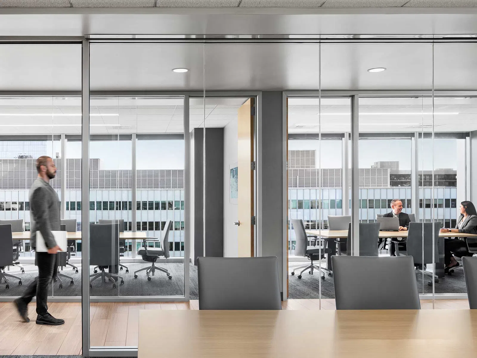 Modern office with glass walls, a man walking, and two colleagues having a meeting with laptops inside a conference room.