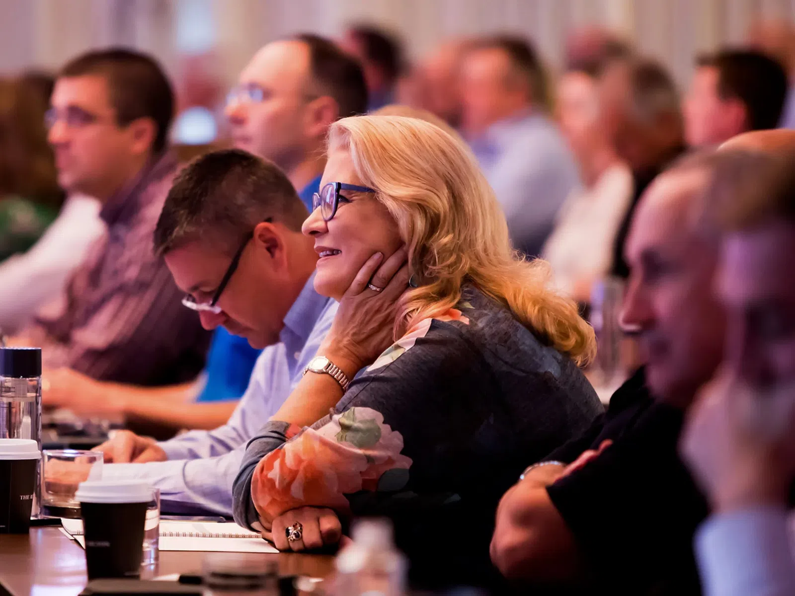 Attentive audience members listening at a conference with notebooks and coffee cups on the table.