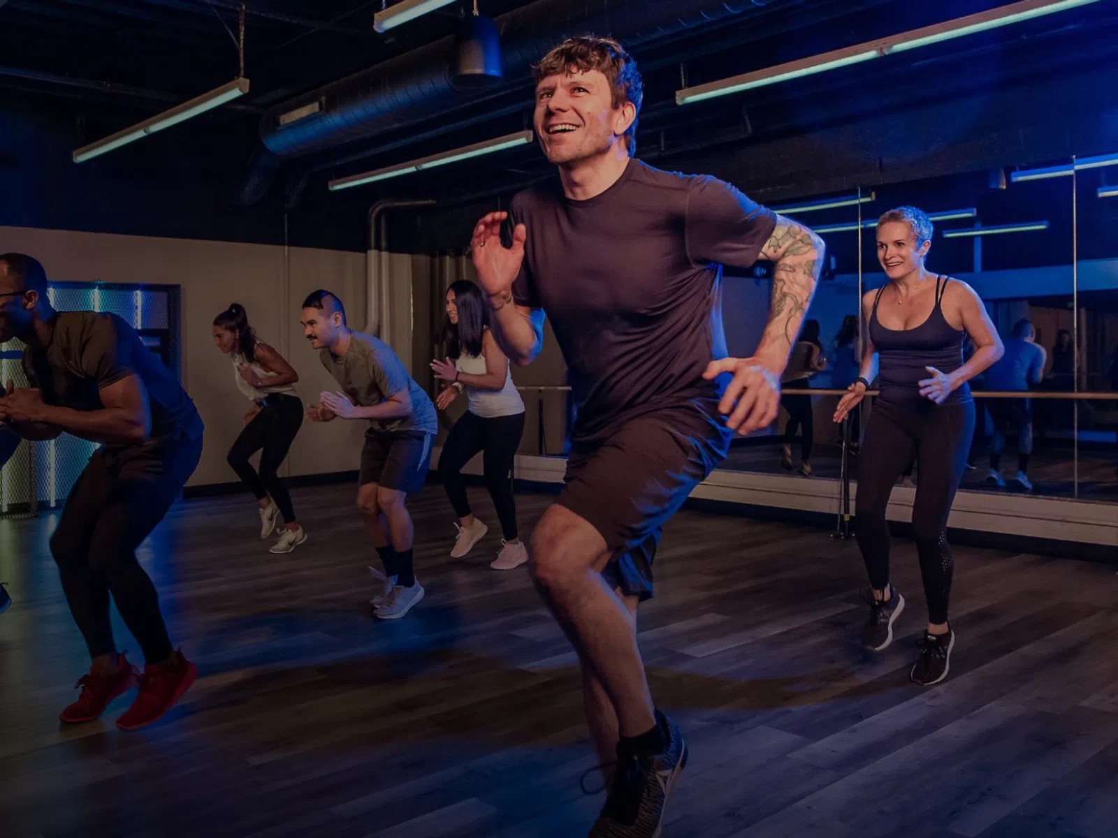 Group fitness class with diverse adults doing aerobic exercises in a dimly lit gym studio with mirrors.
