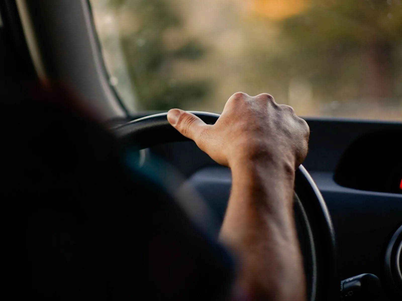 Close-up of a hand gripping a car steering wheel during a drive at sunset with blurred background.