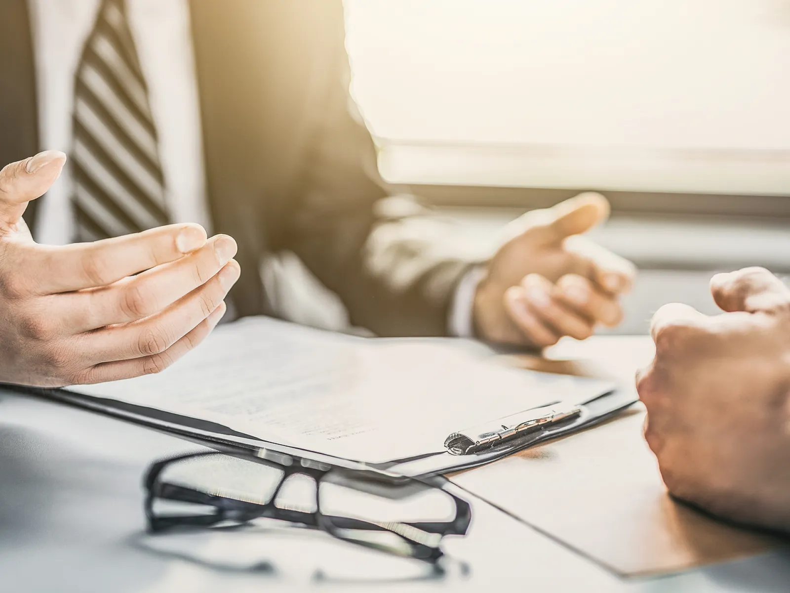 Two professionals discussing documents across a desk with eyeglasses and clipboard in a bright office setting