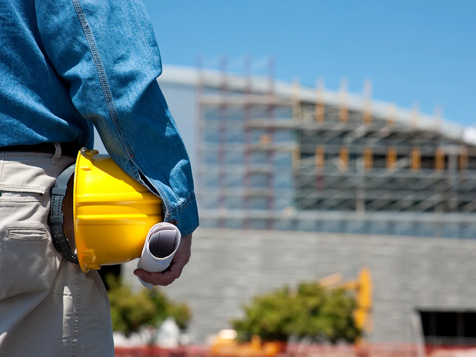 Construction worker holding yellow helmet and blueprints with stadium under construction in the background.