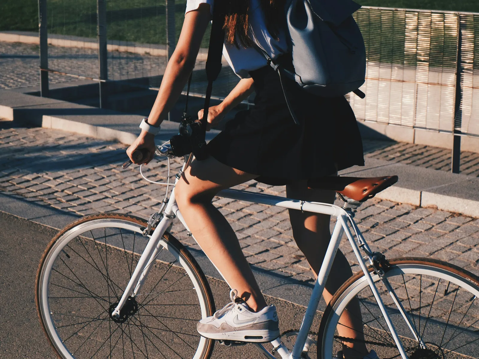 Person riding a white bicycle on a cobblestone street wearing white sneakers and a black skirt