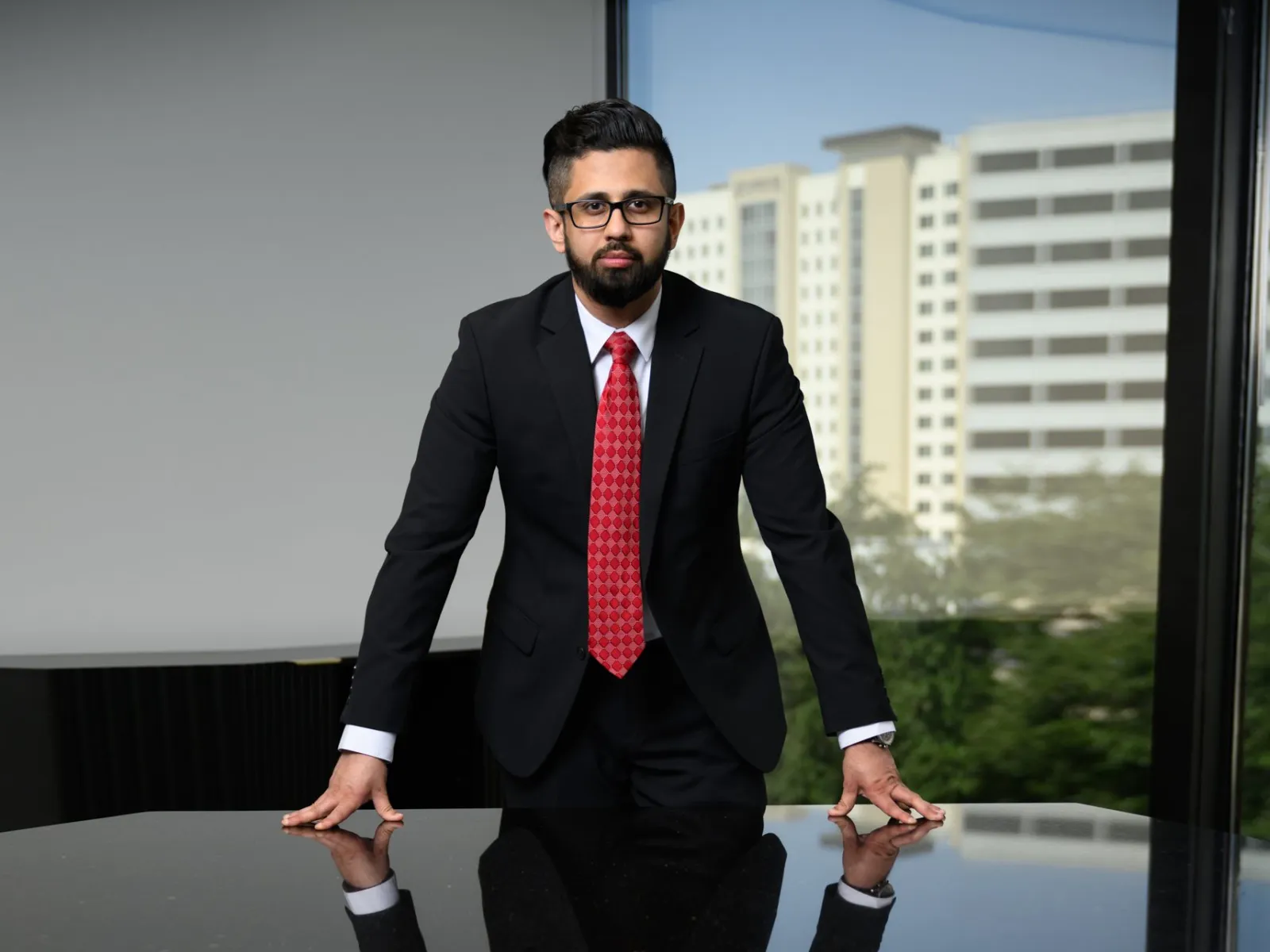 Confident businessman in a black suit and red tie stands with hands on a reflective table in a modern office.