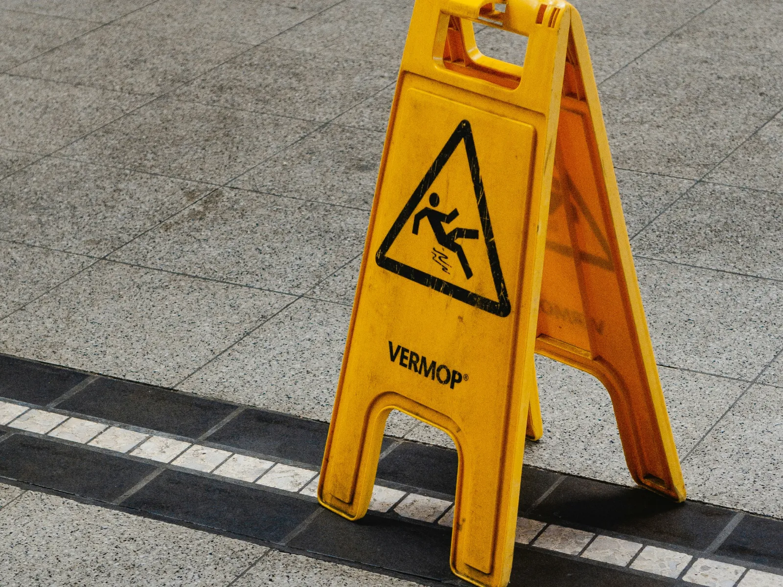 Yellow caution sign warning of slippery floor placed on tiled surface with black and white strip.
