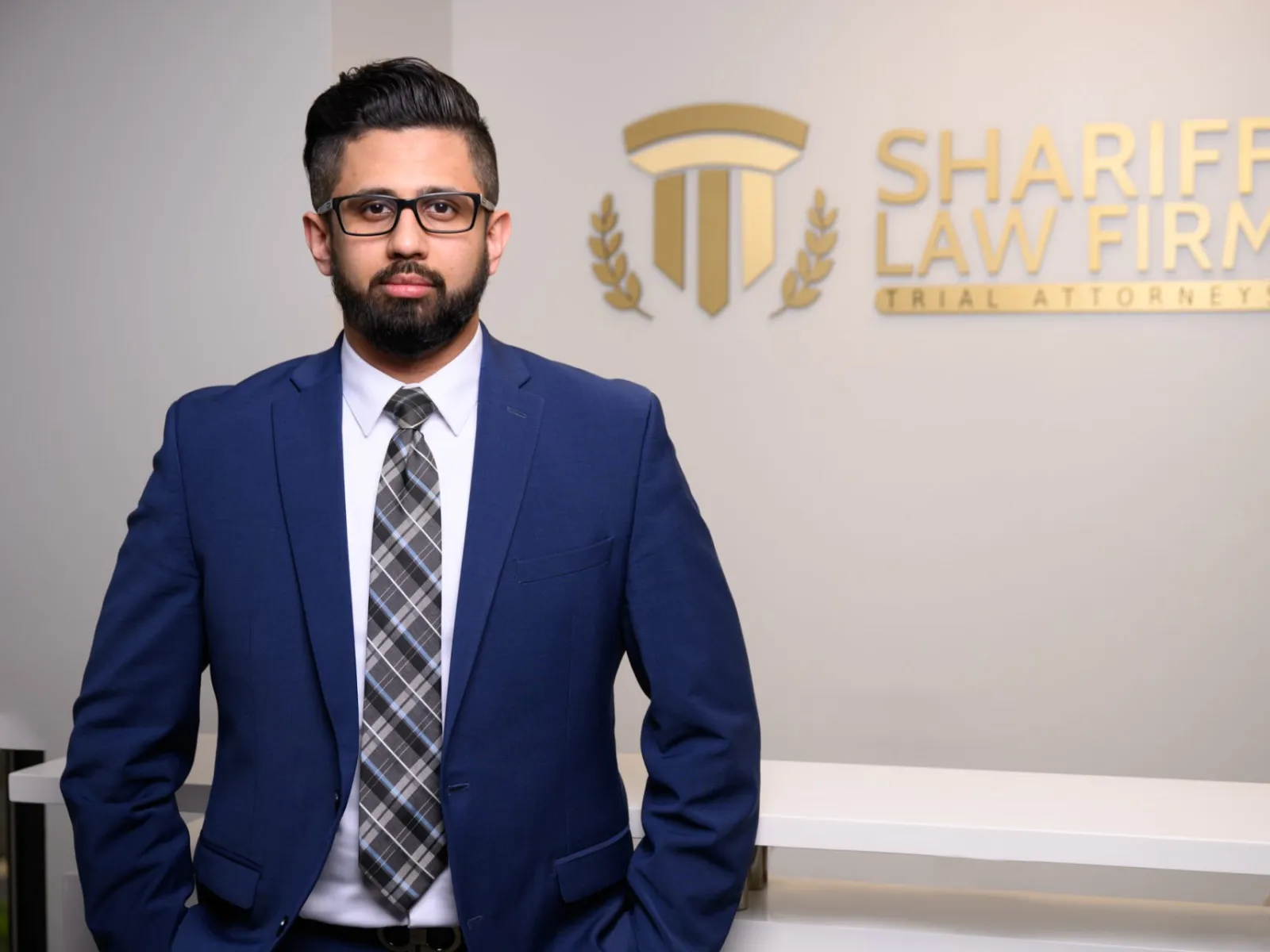 Professional man in blue suit and plaid tie standing in front of Shariff Law Firm logo wall