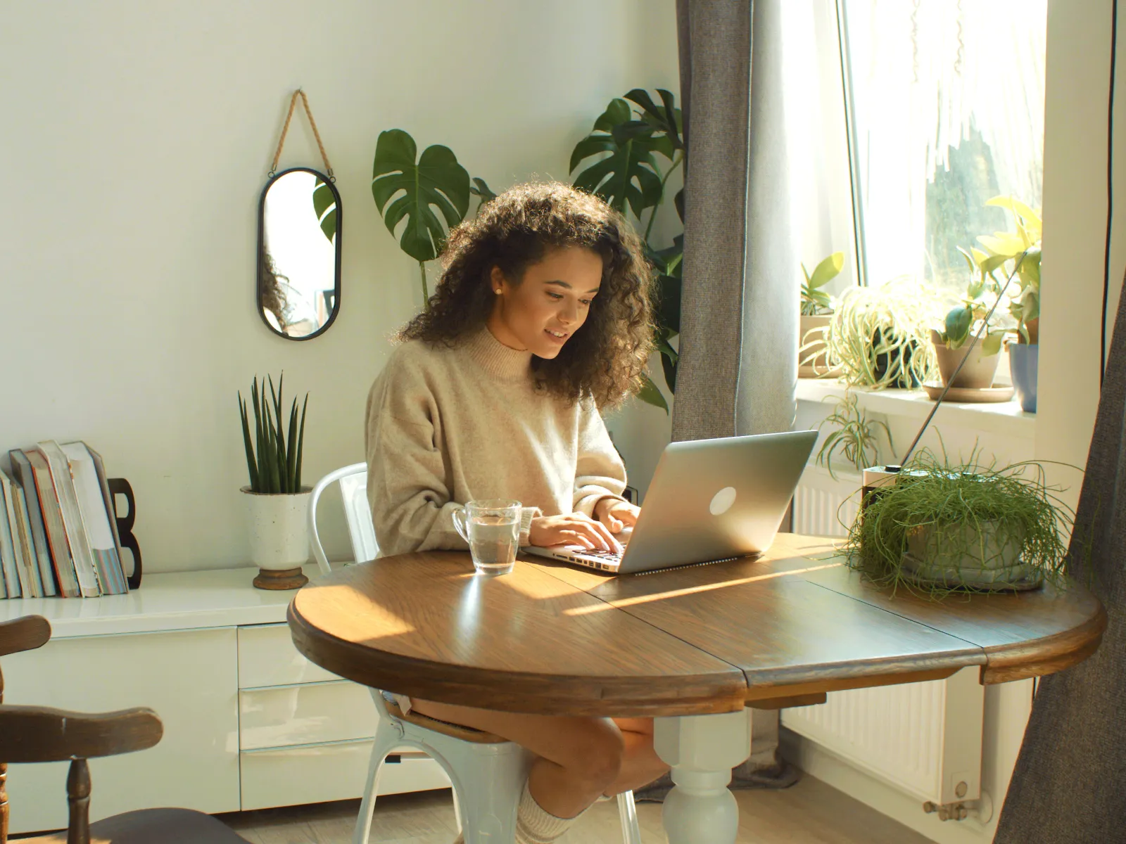 Young woman working on a laptop at a wooden table in a bright, cozy kitchen with plants and natural light.