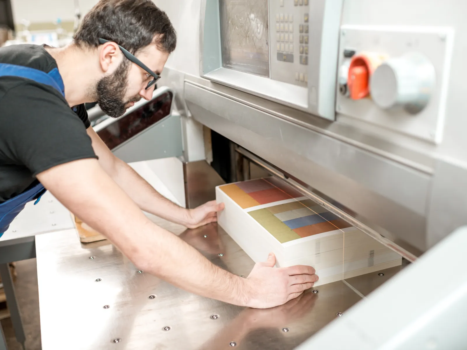 Man operating industrial paper cutting machine handling thick stack of printed sheets with color blocks.
