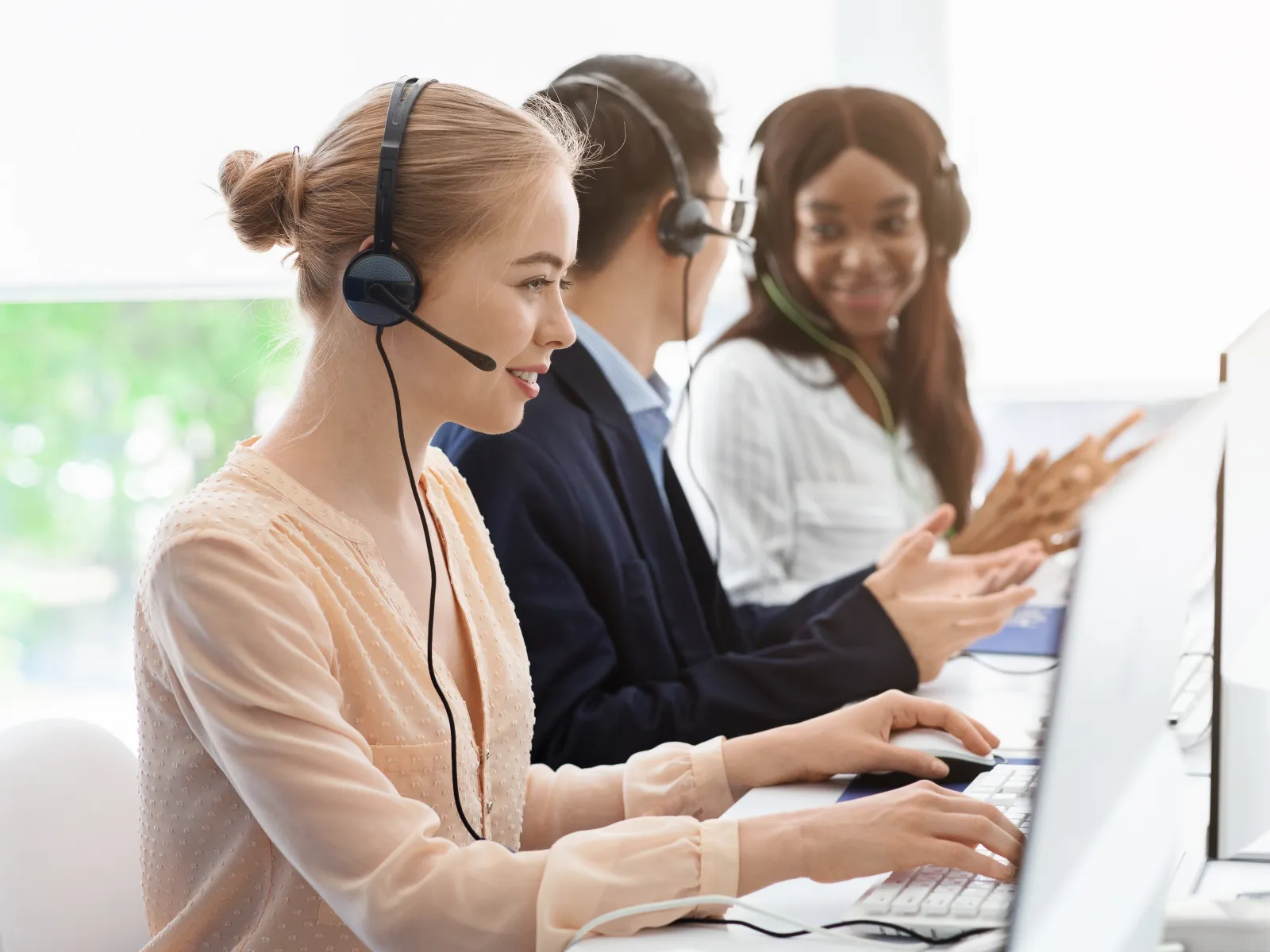 Three diverse customer support agents wearing headsets working at computers and collaborating in office