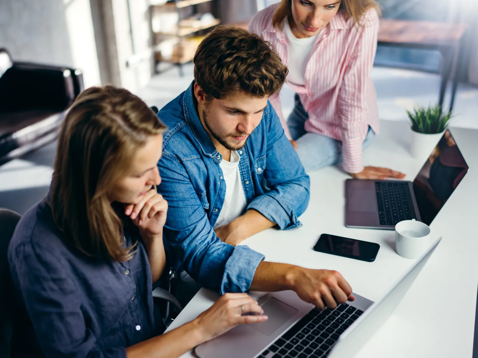 Three young professionals collaborating over a laptop in a modern office space with laptops and coffee.