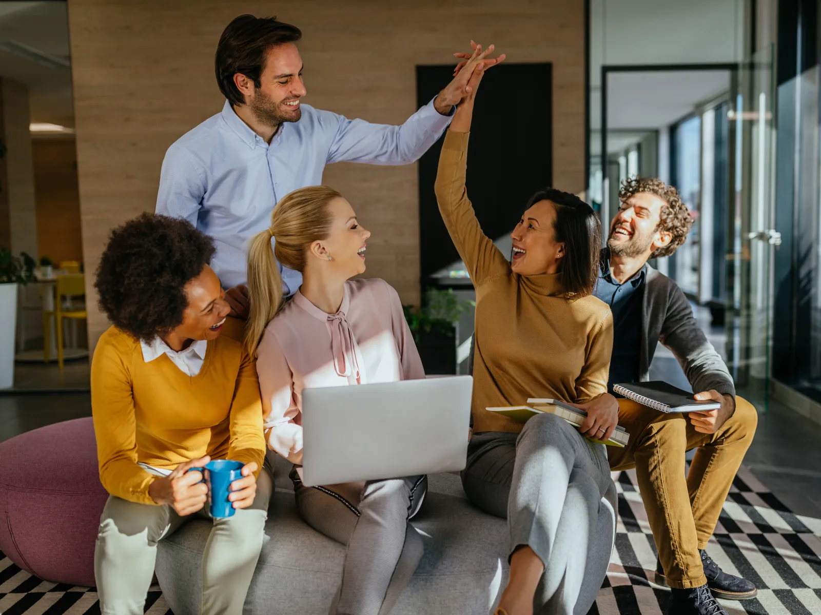 Diverse team of five colleagues collaborating and high-fiving in modern office lounge with laptop and notebooks.