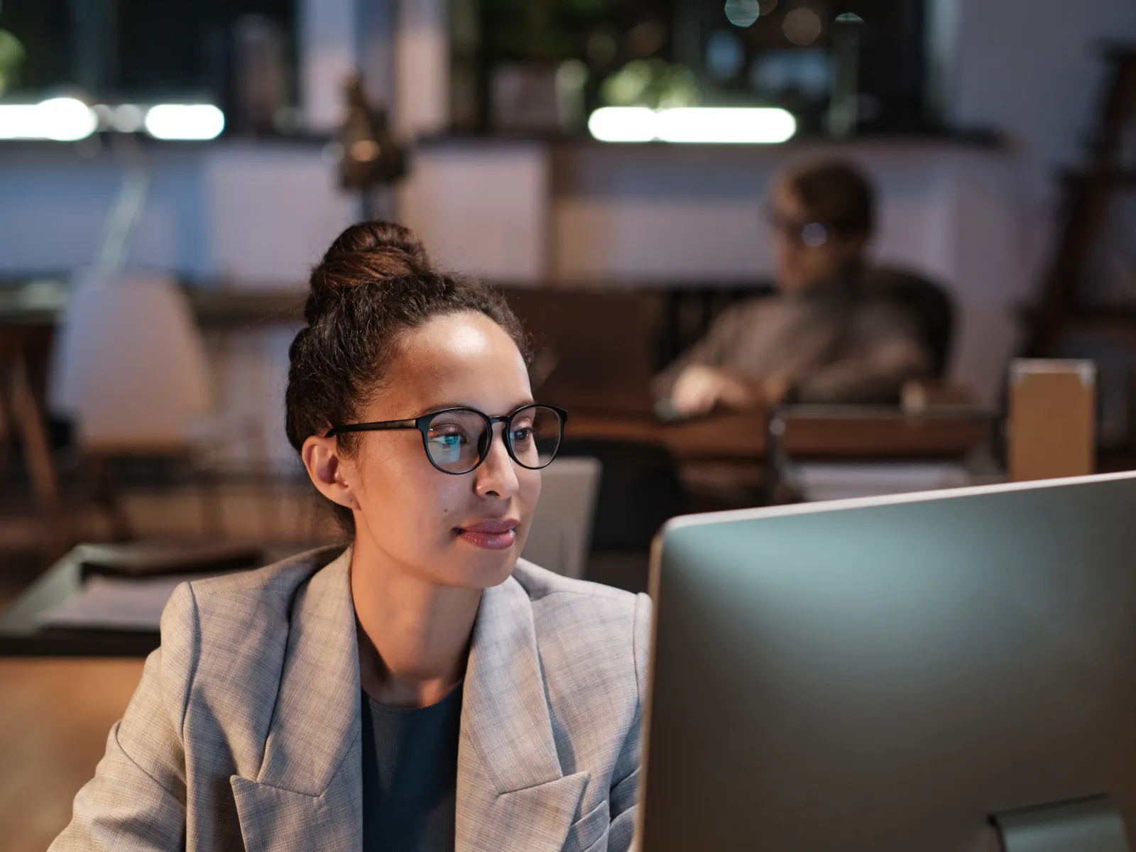 Young woman in glasses working late on a computer in a modern office setting with blurred background.
