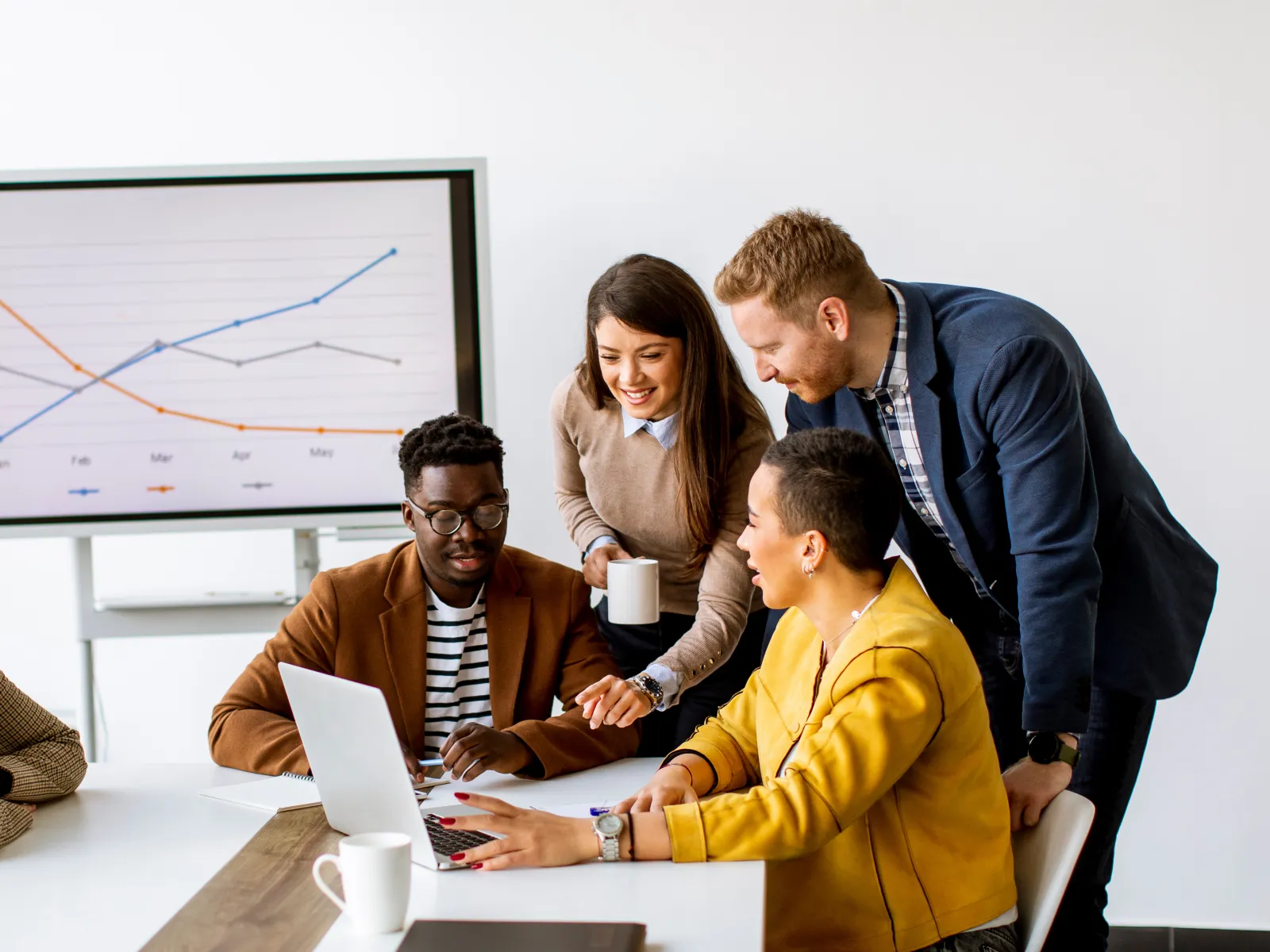 Diverse team gathered around a laptop discussing data charts on a screen in a bright modern office meeting.