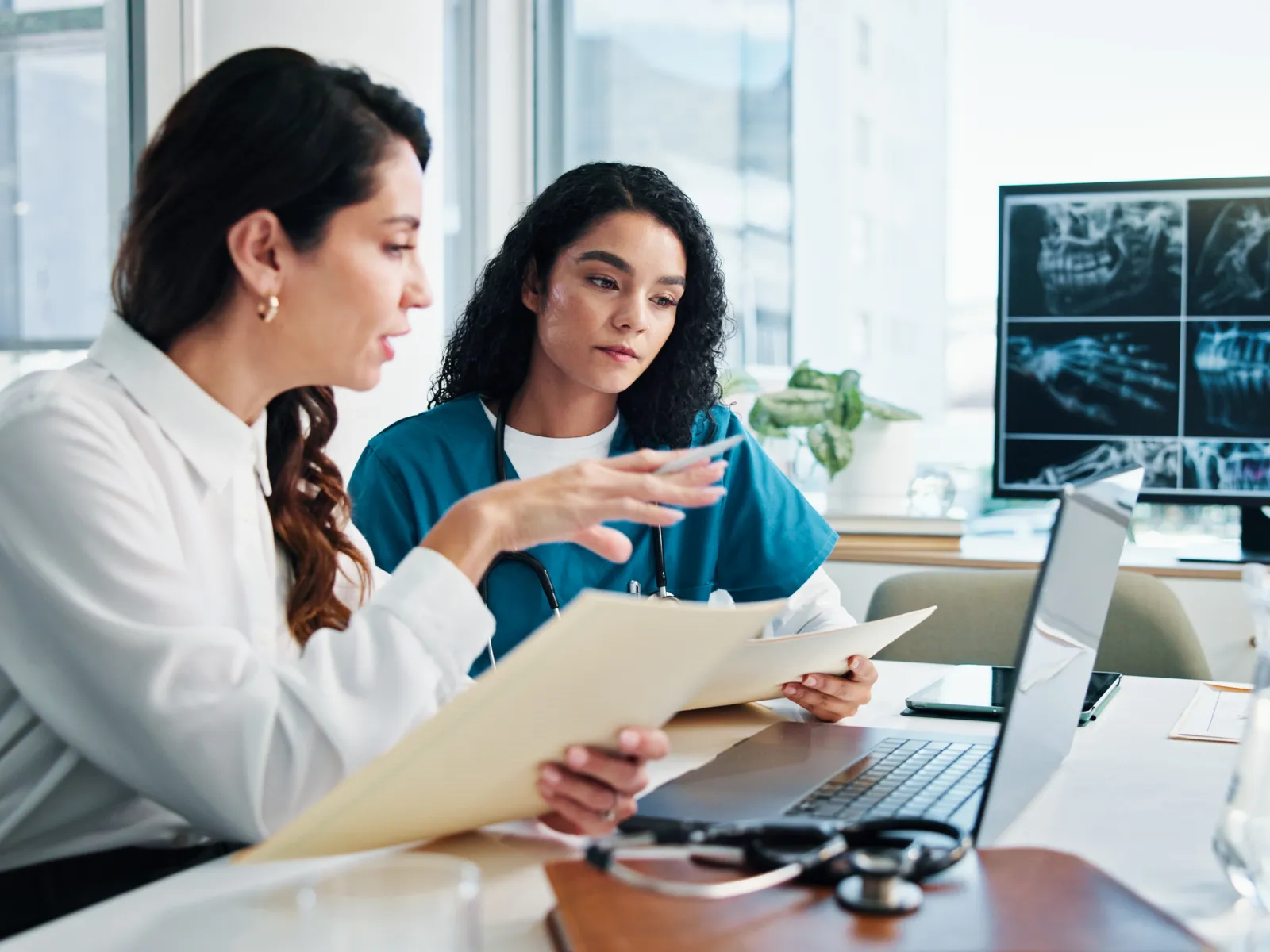 Two female doctors discussing patient files with dental X-rays displayed on a computer monitor.