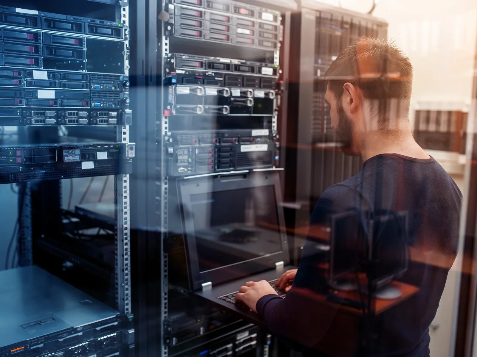 Man working on laptop in a server room surrounded by racks of networking equipment and servers.