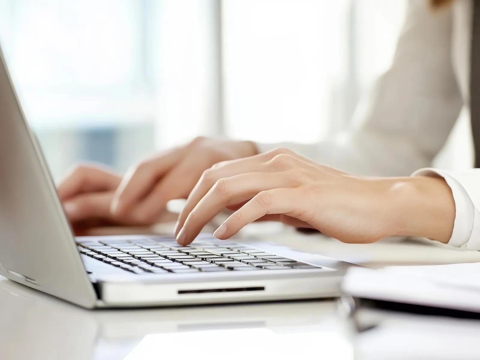 Close-up of hands typing on a laptop keyboard in a bright office setting with documents nearby