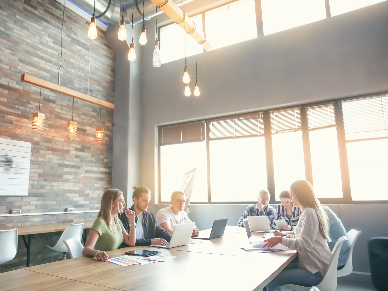 Team of six people working together on laptops and documents in a bright modern office with brick walls.