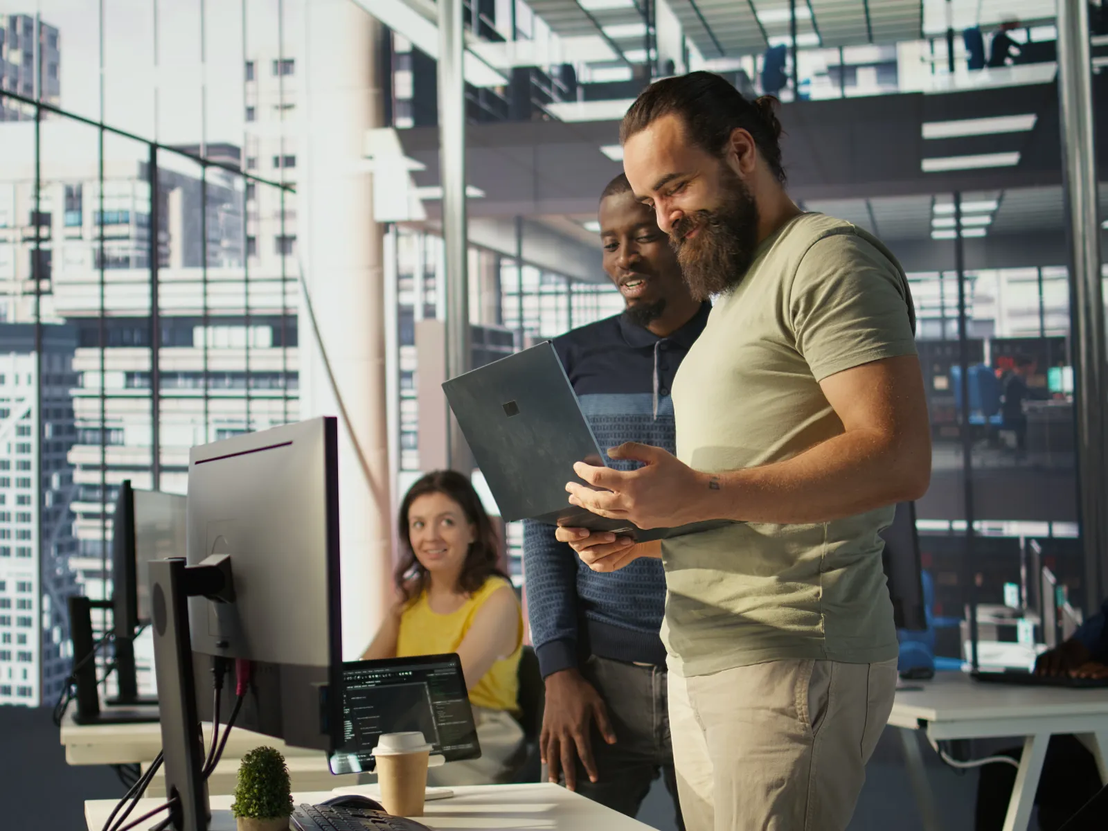 Two men discussing work using a laptop while a woman works at a desktop in a modern office with city views.