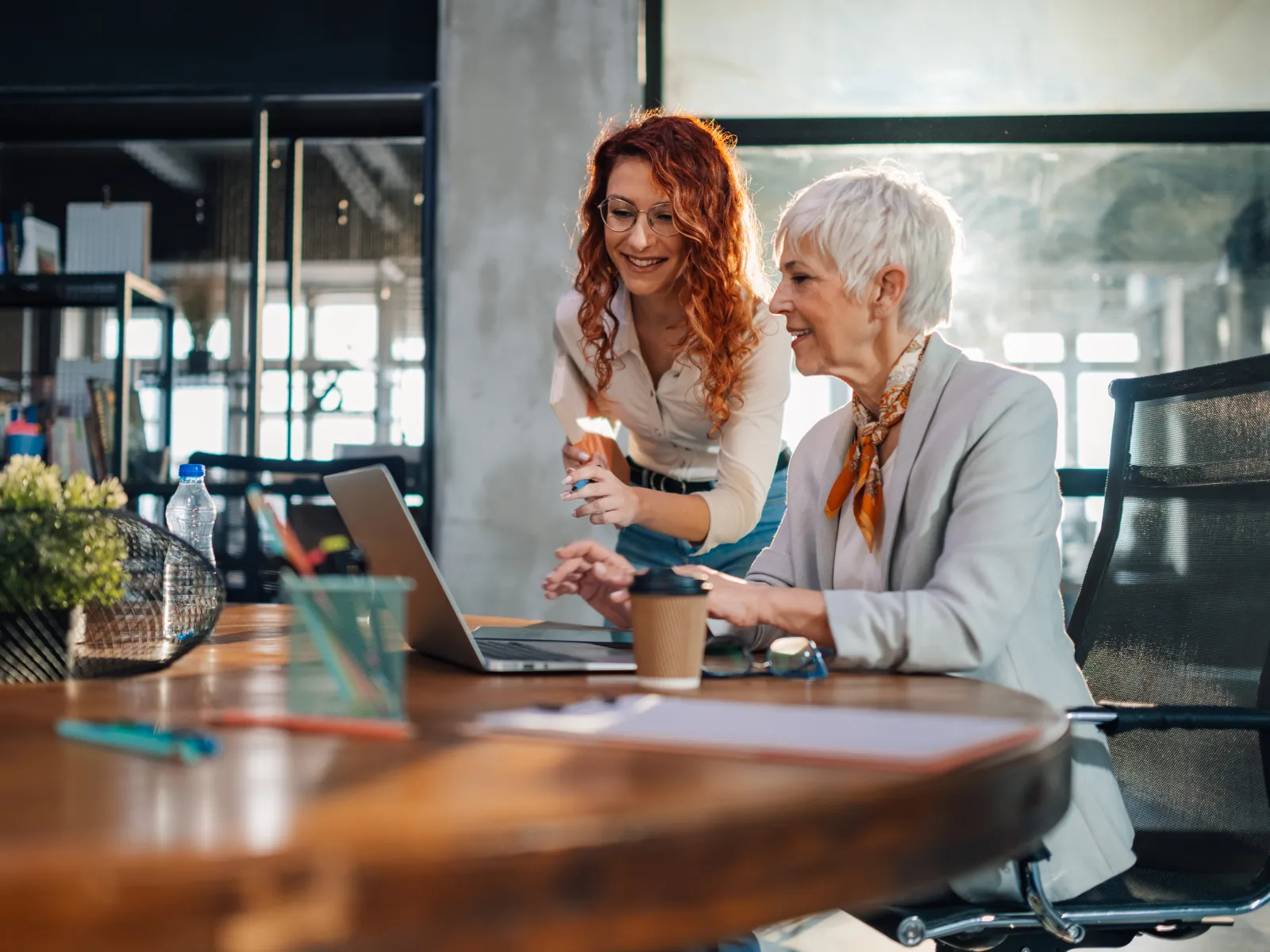 Two professional women collaborate on a laptop in a modern office with natural light and wooden table.