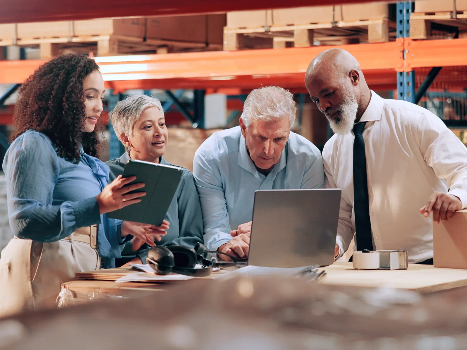 Diverse team gathered around laptop in warehouse discussing work and logistics with packages and shelves in background