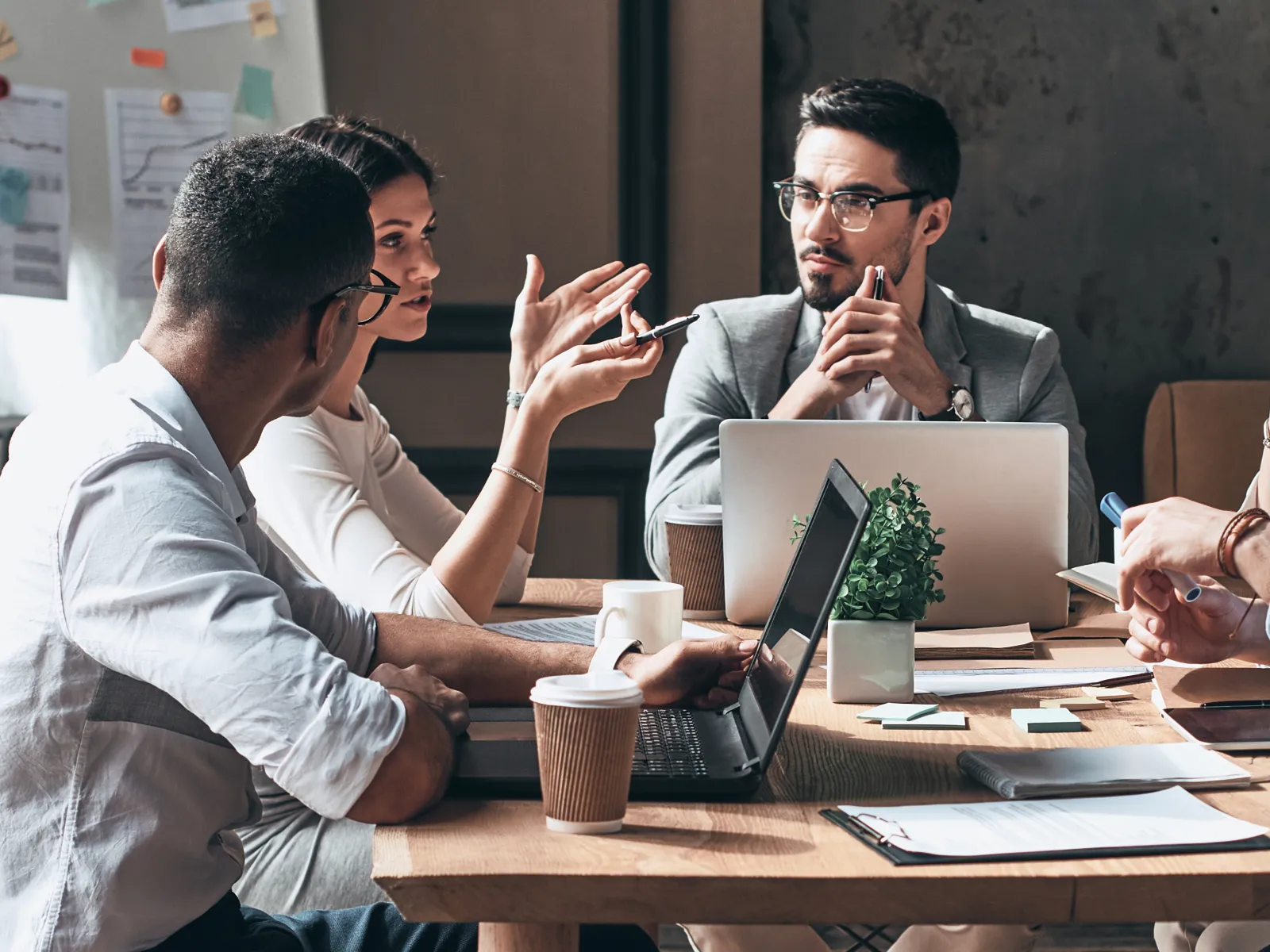 Five diverse professionals engaged in a meeting around a wooden table with laptops and coffee cups in a modern office.