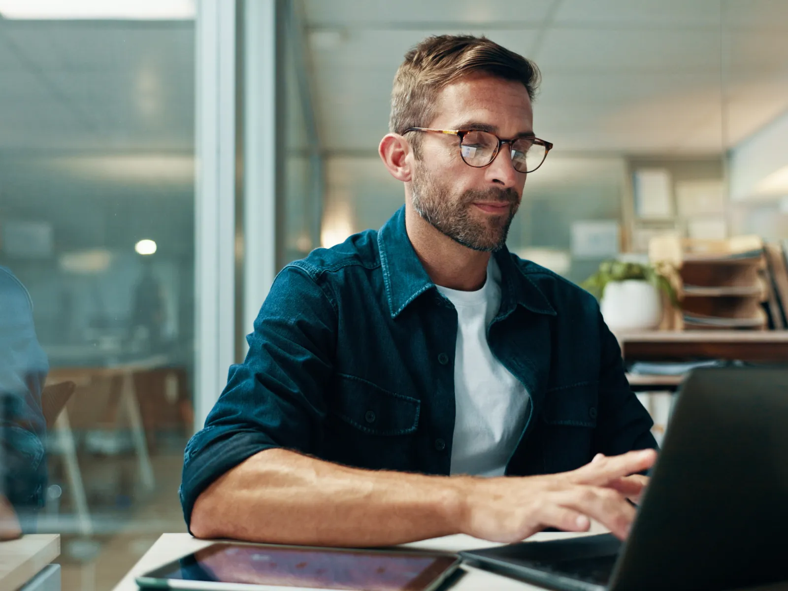 Man wearing glasses working focused on laptop at modern office desk with digital tablet and shelves in background