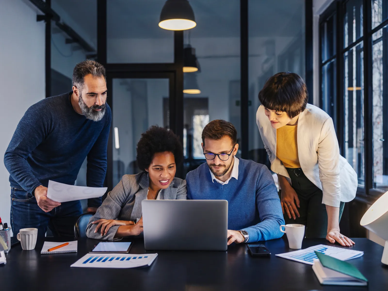 Four diverse professionals collaborating over laptop and reports in modern office with large windows and pendant lights.