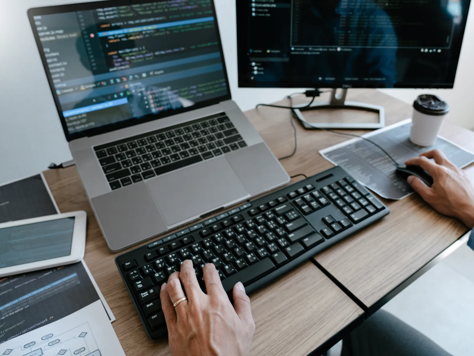 Developer's hands coding software on laptop and desktop with keyboard, mouse, tablet, and coffee on wooden desk.