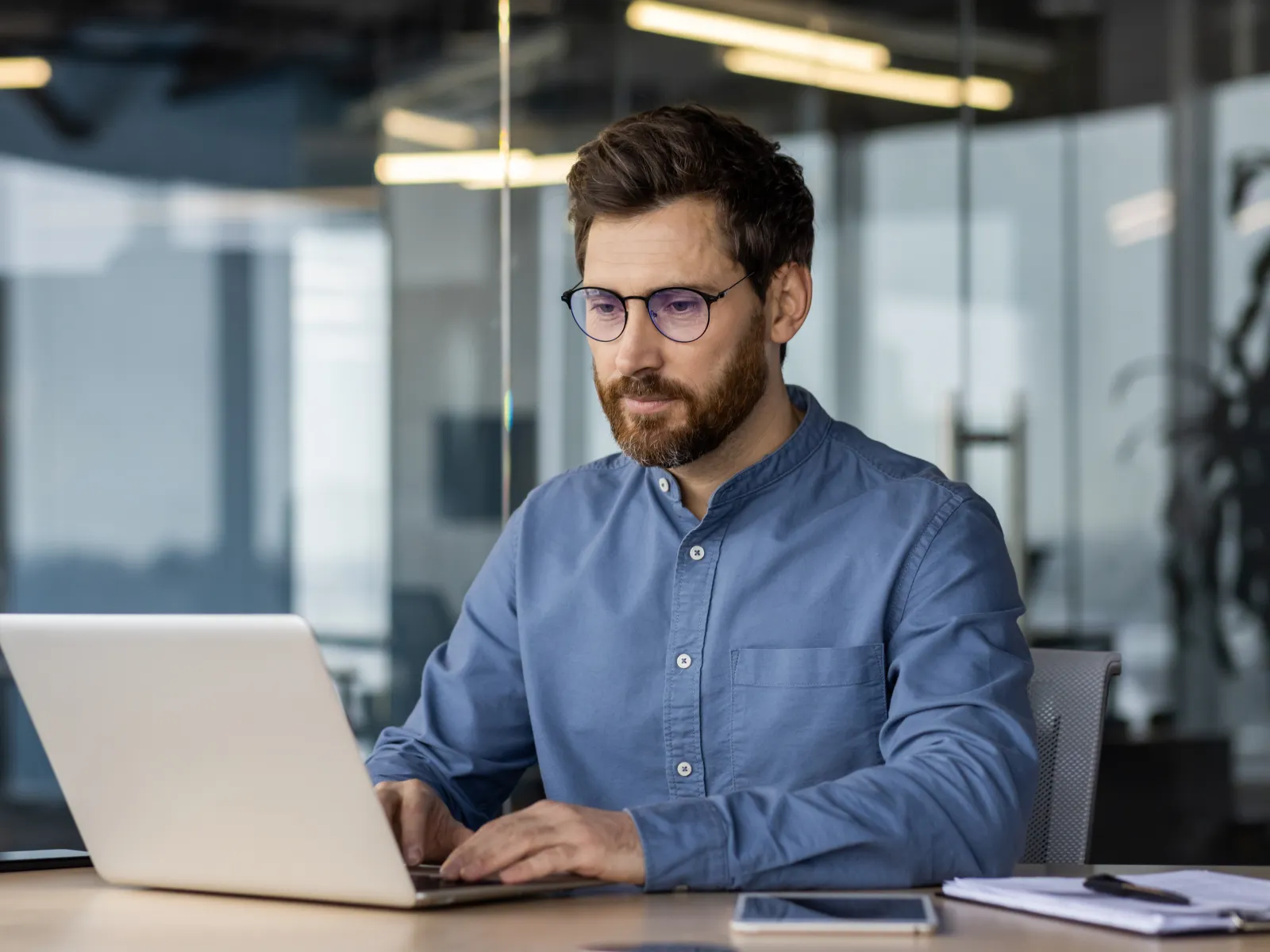 Man wearing glasses working on a laptop at a desk in a modern office environment focused on his task