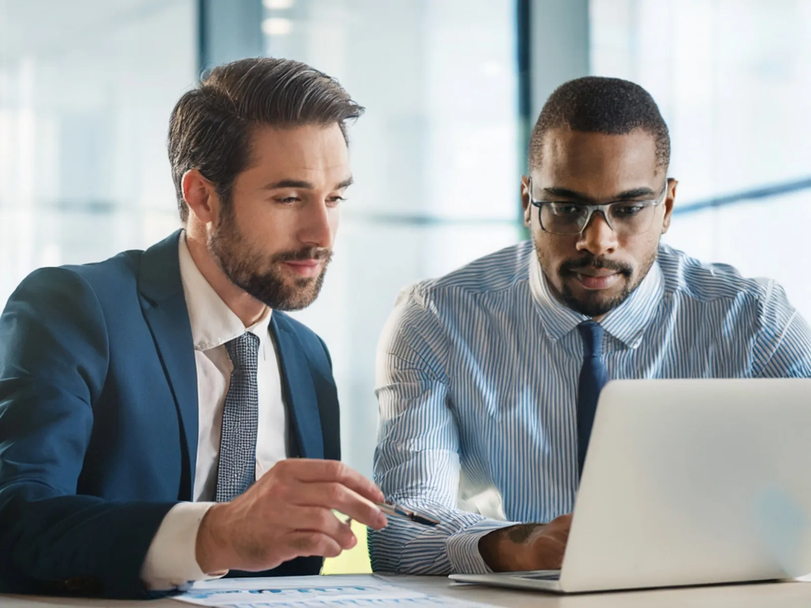 Two businessmen in suits working together on a laptop in a bright modern office setting.
