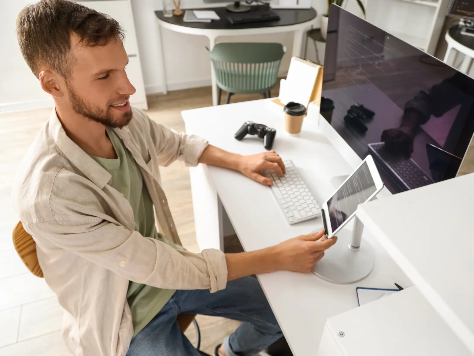 Man working at desk with computer, tablet, keyboard, coffee cup, and game controller in bright modern office space
