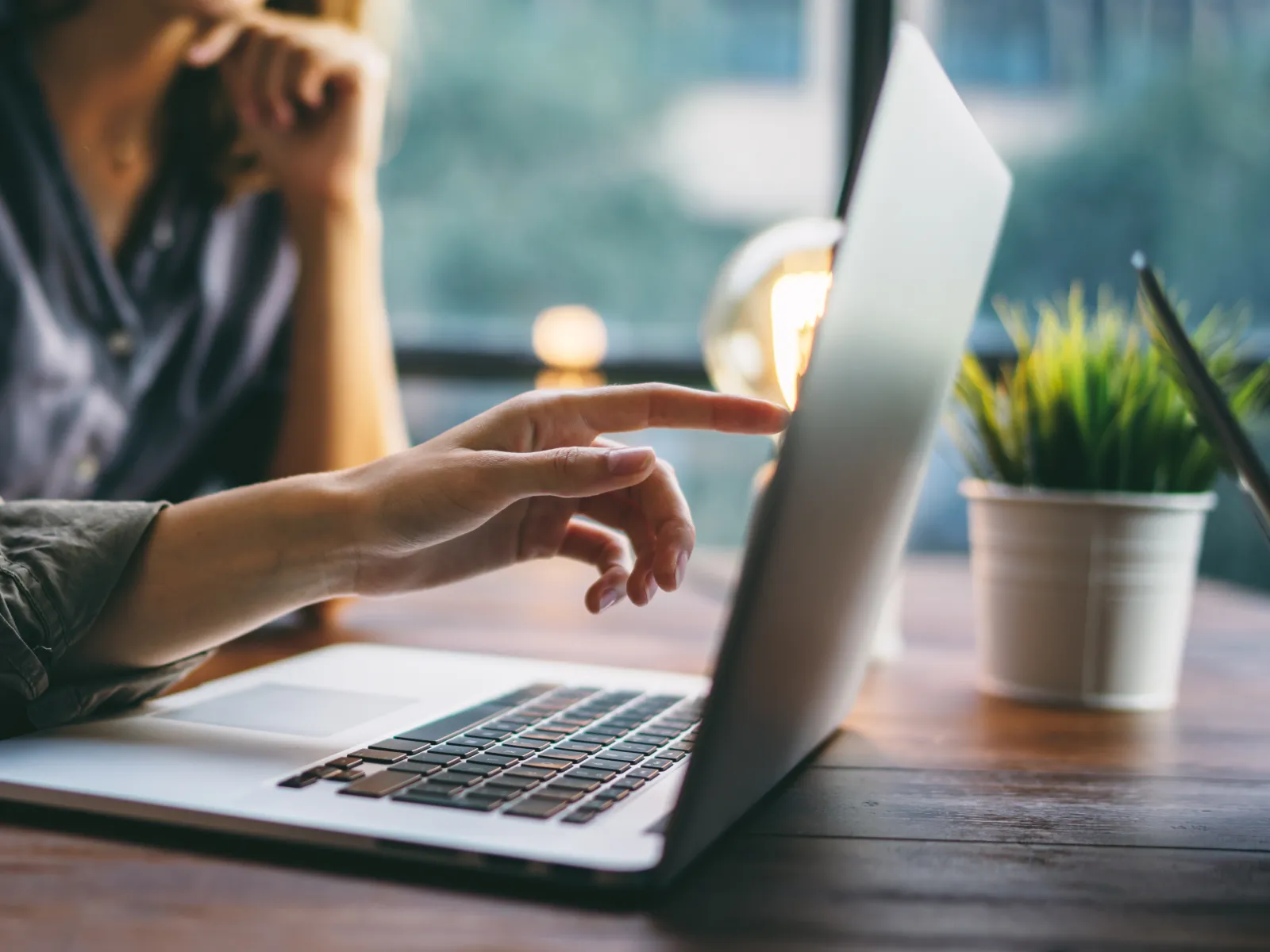 Person pointing at laptop screen with another person in background discussing work in bright office