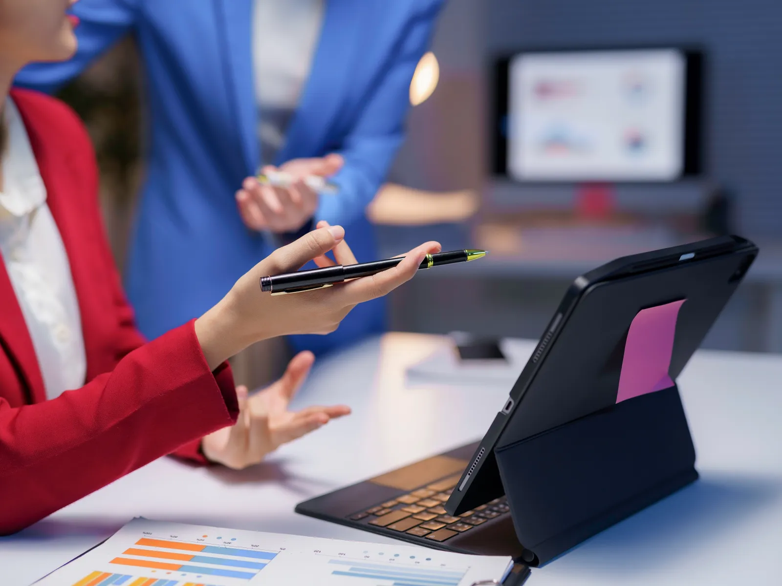 Two businesswomen discussing work with charts and a tablet on a desk in an office setting.