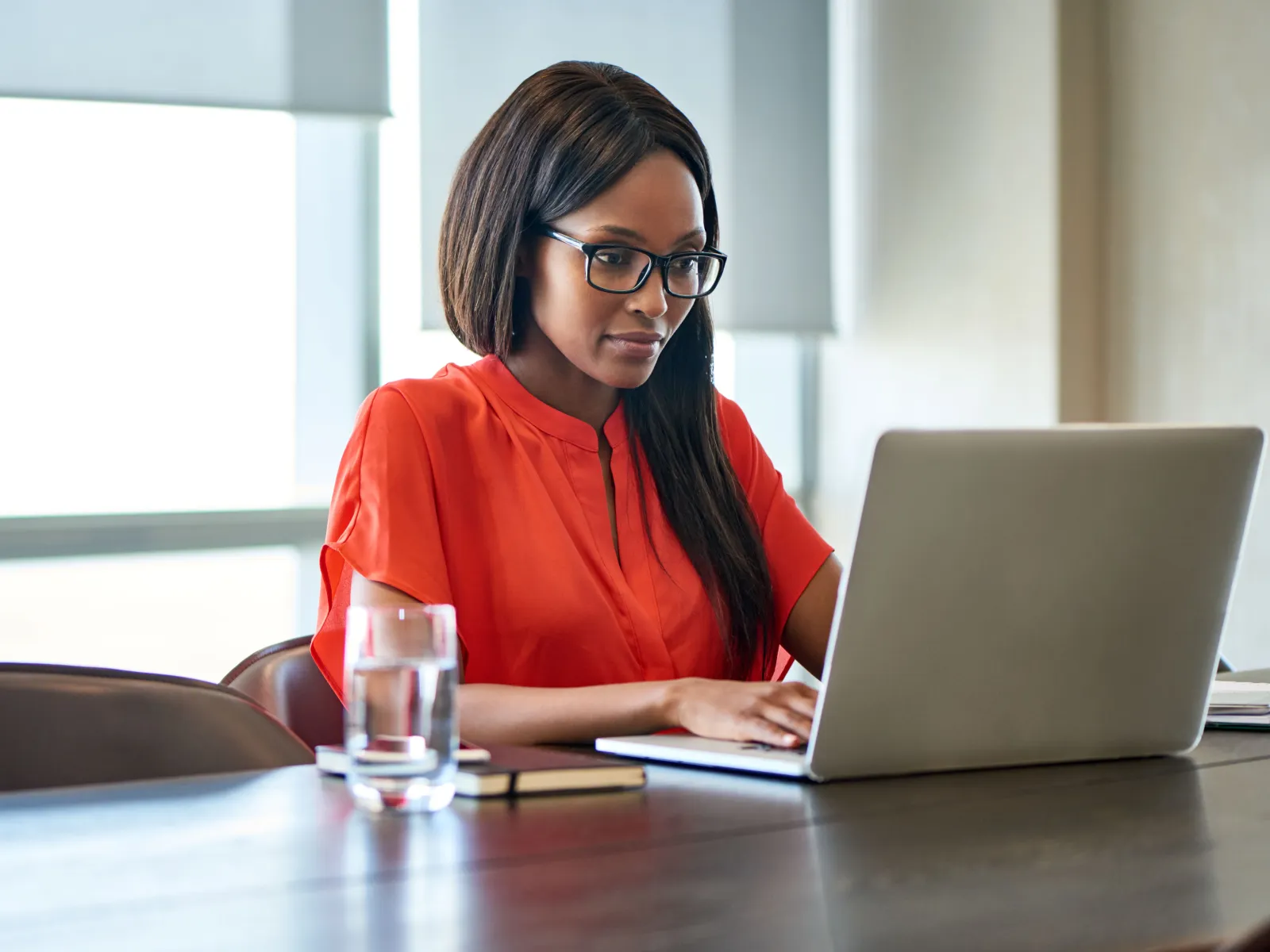 Woman in red blouse working on laptop at office table with glass of water and notebook nearby.