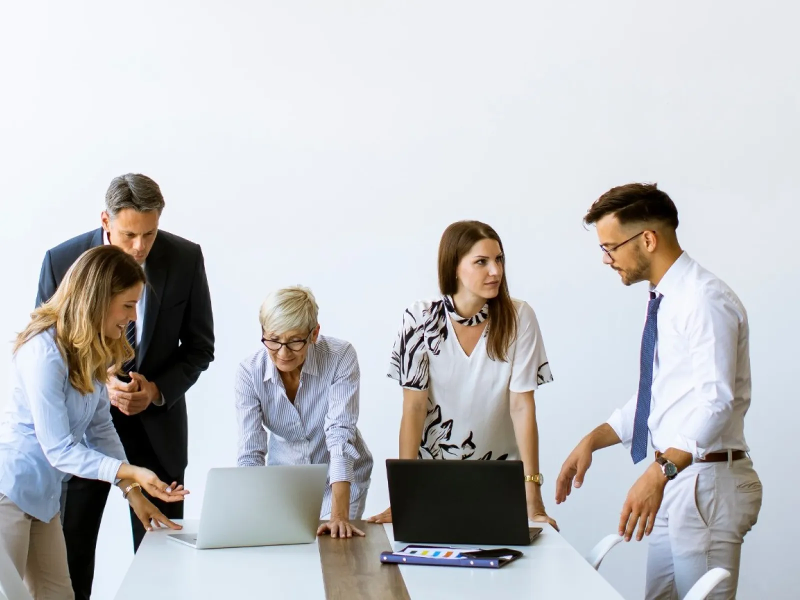 Five professionals collaborating around a table with laptops in a bright office with white walls