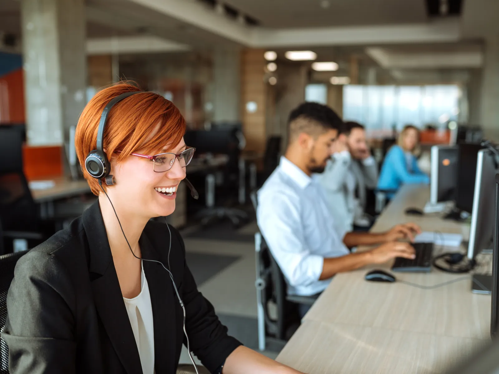 Smiling female customer service agent wearing headset working at computer with colleagues in modern office