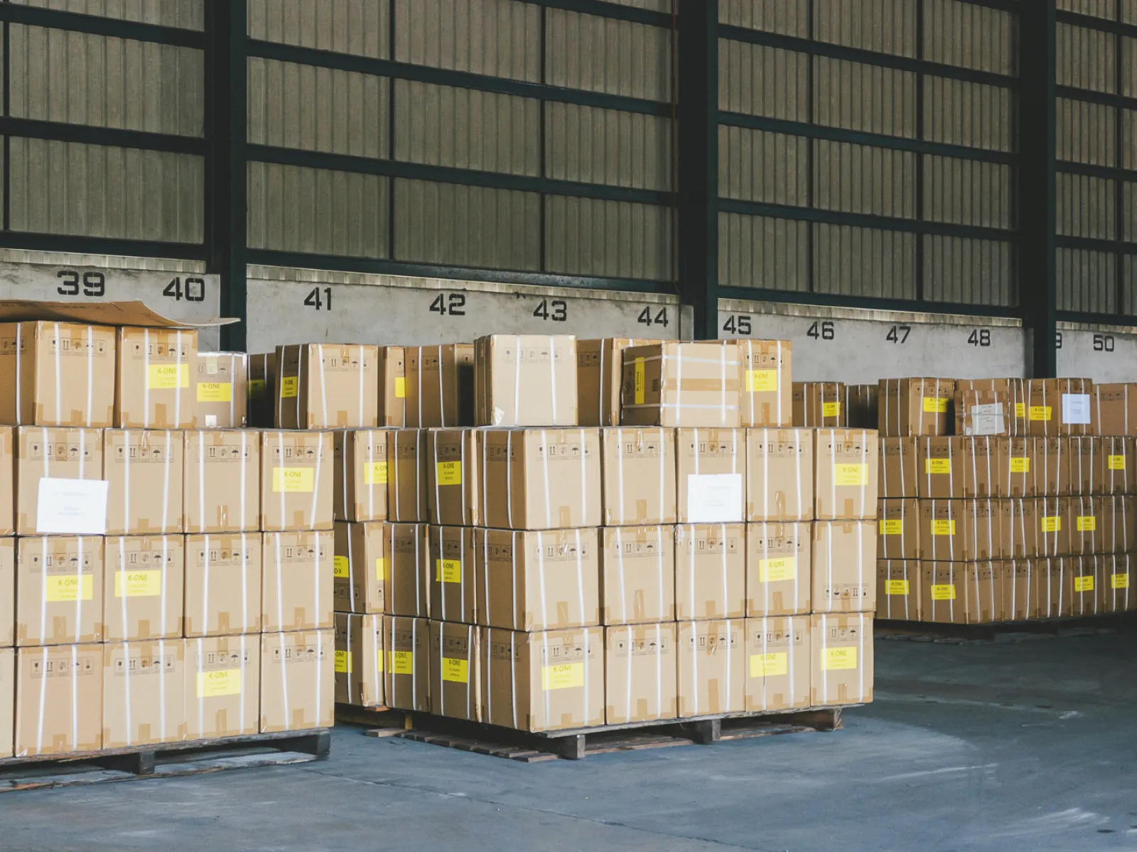 Rows of stacked cardboard boxes on pallets inside a spacious warehouse with numbered loading bays.