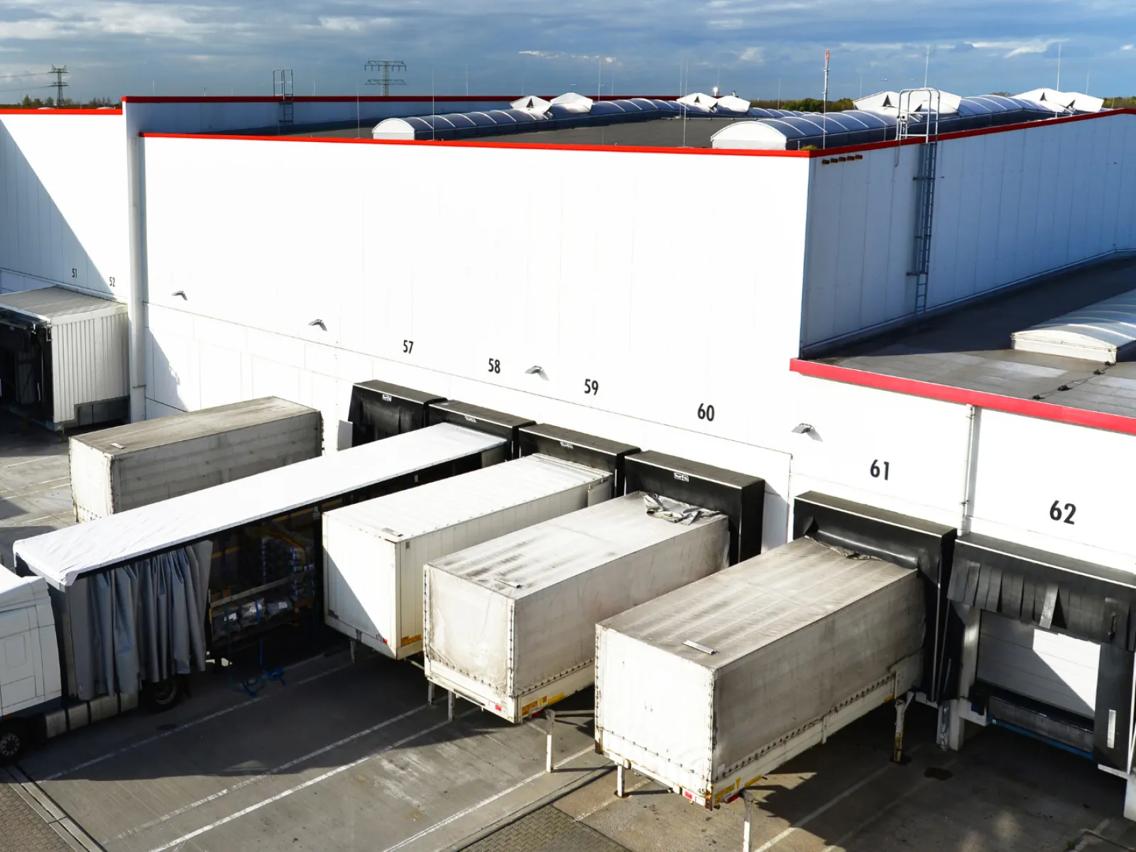 Large warehouse loading dock with multiple trucks backed into numbered bays under a partly cloudy sky