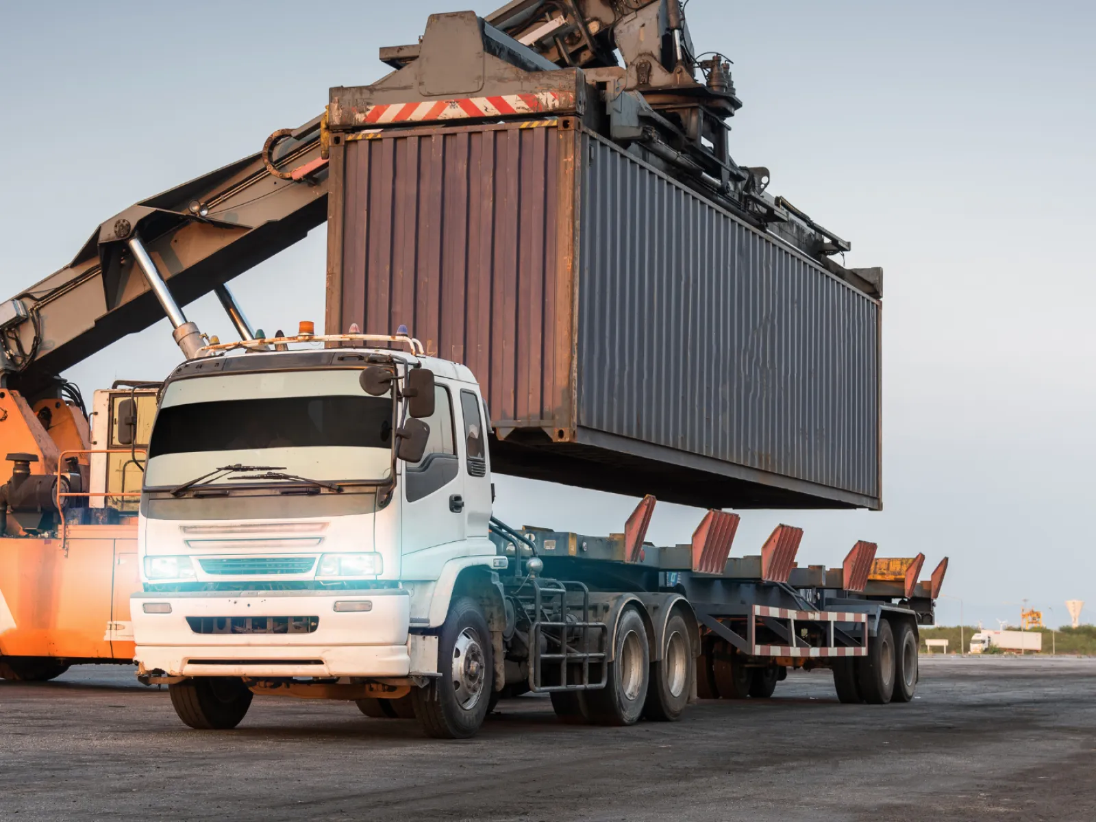 White flatbed truck while a large orange crane loads a shipping container onto it at a port during dusk.