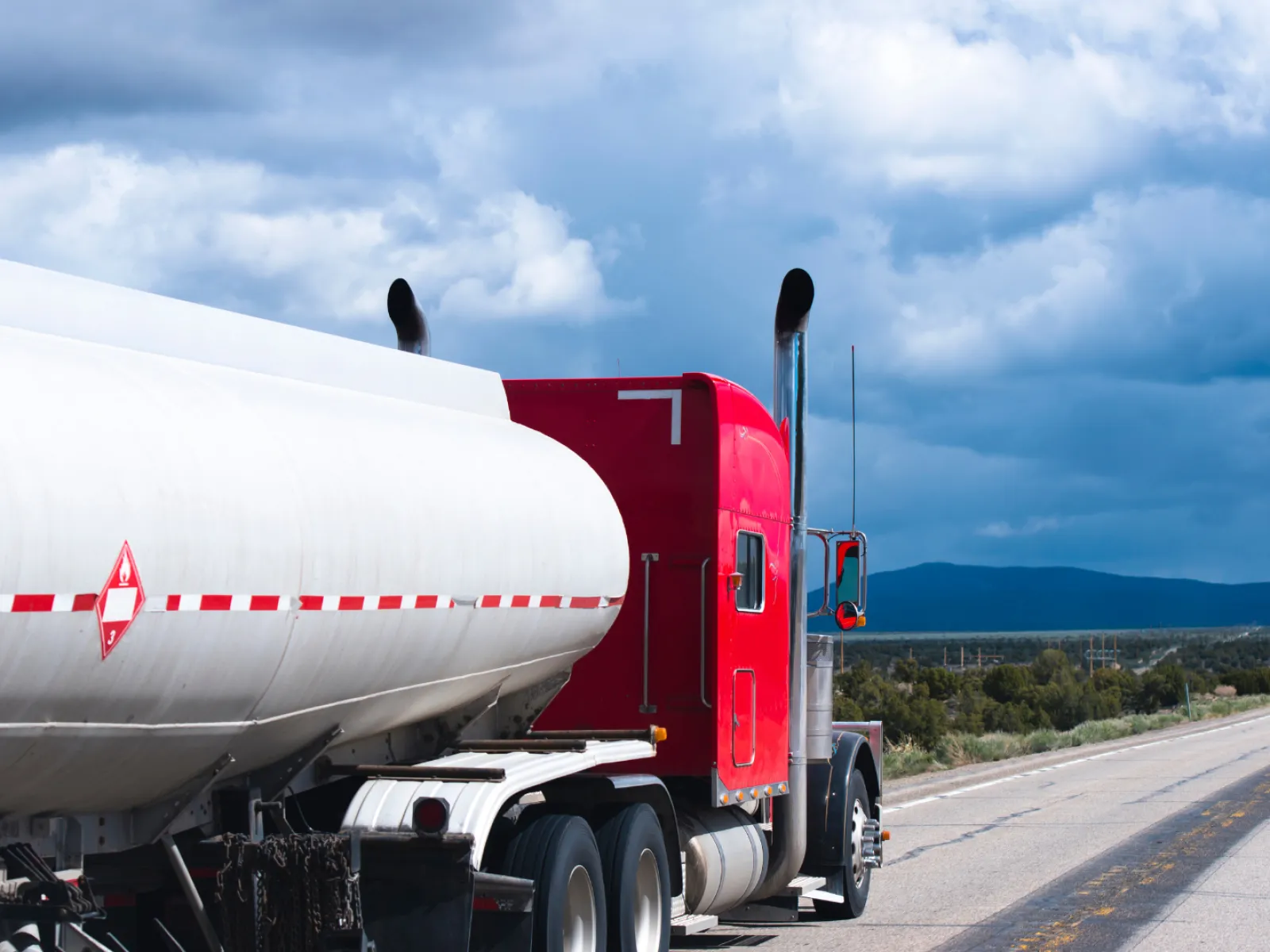 Red tanker truck driving on an open highway under a cloudy sky with mountains in the distance
