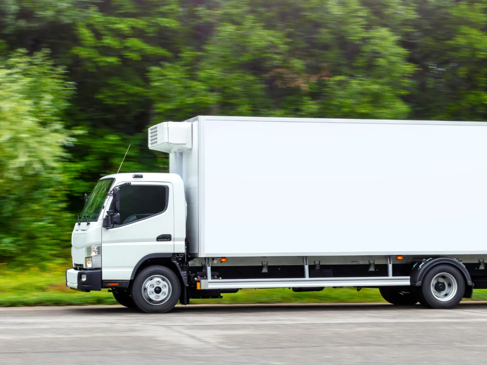 White refrigerated box truck driving on road with green trees in background on a clear day